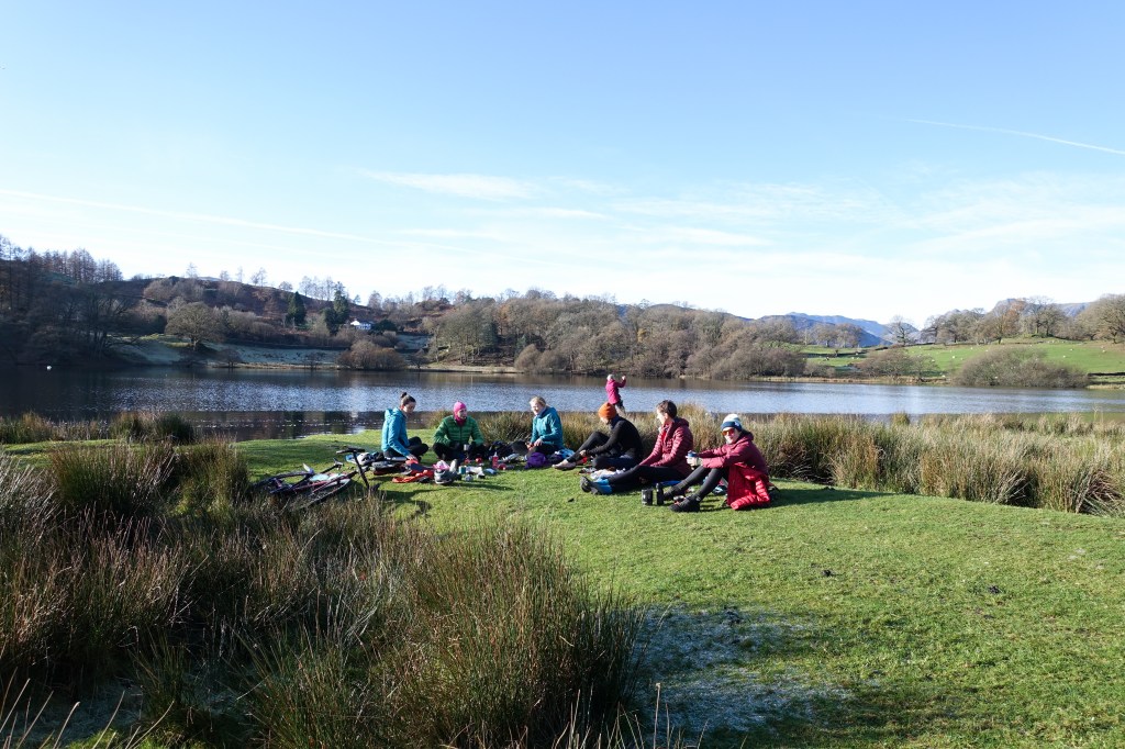 group of women sat by a tarn drinking hot chocolate mid-bike ride